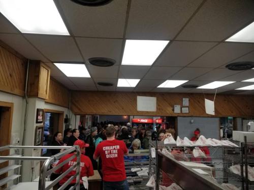 A crowded restaurant with people waiting in line, staff in red shirts, and a cozy wooden interior.