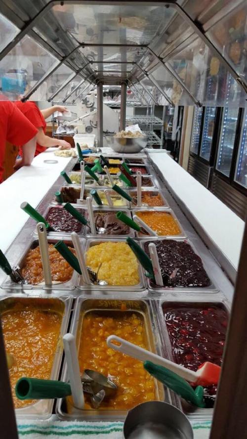 A variety of colorful fruit toppings in metal trays, with green serving spoons, displayed in a dessert shop.