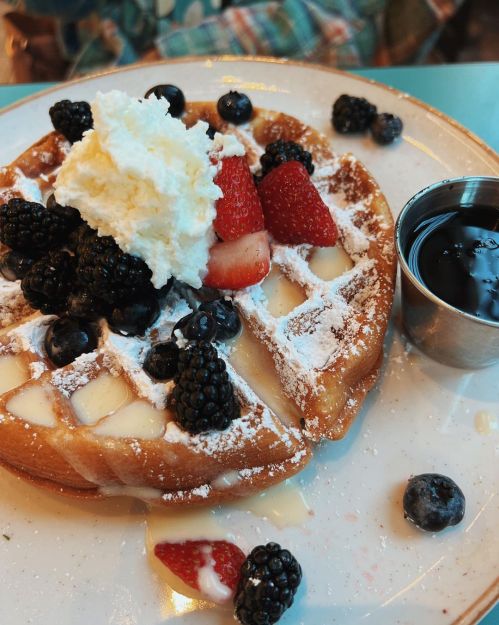 A plate of waffles topped with whipped cream, strawberries, blackberries, and a side of syrup.