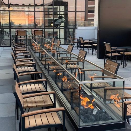 A modern outdoor seating area with a long glass fire feature and wooden chairs, set against a city skyline.
