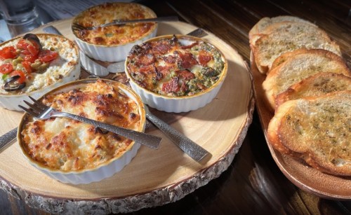 A wooden platter with four baked dishes and toasted bread slices on the side, garnished with herbs.
