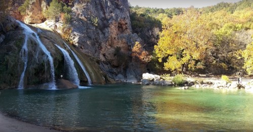 A serene waterfall cascades into a clear pool, surrounded by autumn foliage and rocky cliffs.