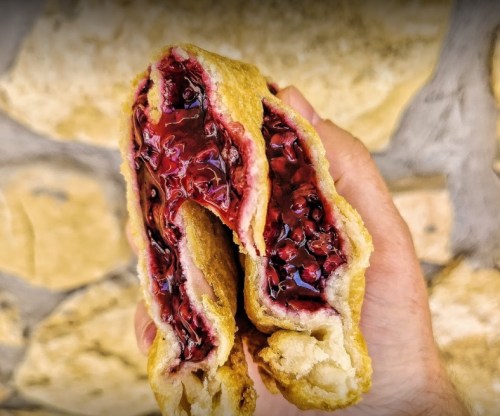 A hand holding a split pastry filled with dark red fruit filling, against a stone wall background.