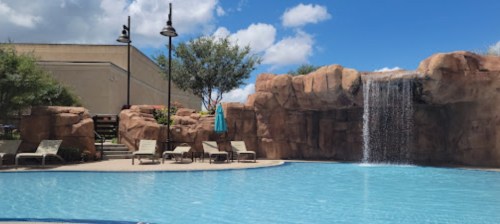 A serene pool area with lounge chairs, a waterfall, and blue skies with fluffy clouds in the background.