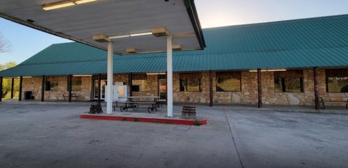 A gas station with a green metal roof and stone walls, featuring a covered pump area and empty parking lot.