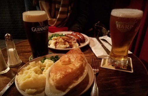 A table with two pints of beer and plates of food, including mashed potatoes, peas, and a meat pie.