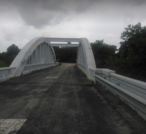 A white arch bridge spans a road, surrounded by greenery under a cloudy sky. The pavement shows signs of wear.