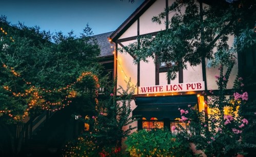 A charming pub with a sign reading "White Lion Pub," surrounded by greenery and warm string lights at dusk.