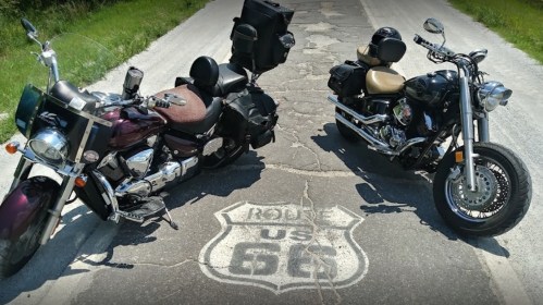 Two motorcycles parked on a cracked road with a "Route 66" sign painted on the pavement. Green grass in the background.