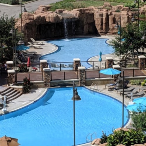 A view of two swimming pools surrounded by lounge chairs, umbrellas, and a waterfall in a landscaped area.