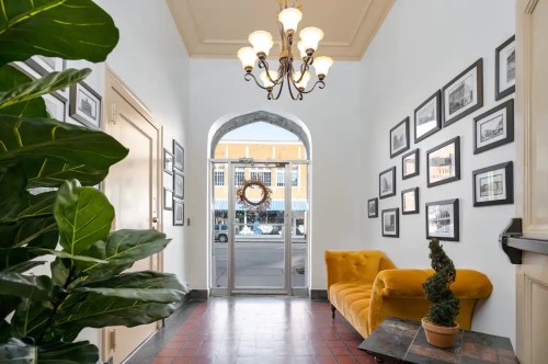 A bright hallway with a yellow couch, potted plants, and framed photos on the walls, leading to a large arched doorway.