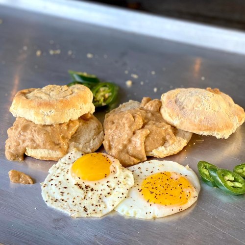 Two biscuits with creamy spread, accompanied by two sunny-side-up eggs and sliced jalapeños on a metal surface.