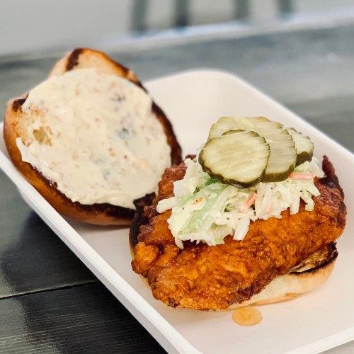 A crispy fried chicken sandwich with pickles and slaw on a toasted bun, served on a white plate.