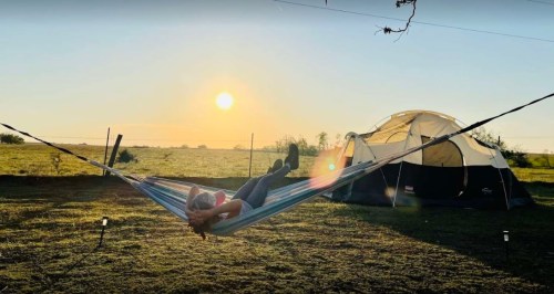 A person relaxes in a hammock at sunset, with a tent set up nearby in a grassy field.