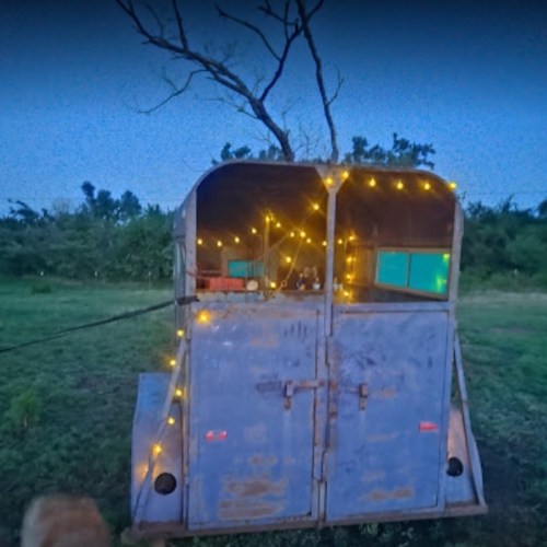 A rustic trailer adorned with string lights, set in a grassy area under a twilight sky.