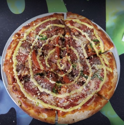 A circular pizza topped with various meats, vegetables, and a spiral of yellow sauce, served on a silver tray.