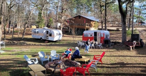 A cozy outdoor scene with two vintage trailers, a fire pit, and people sitting in colorful chairs among trees.
