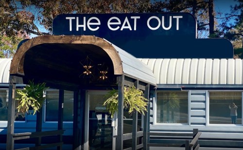 Sign for "The Eat Out" restaurant with a wooden entrance and hanging plants. Bright blue sky in the background.