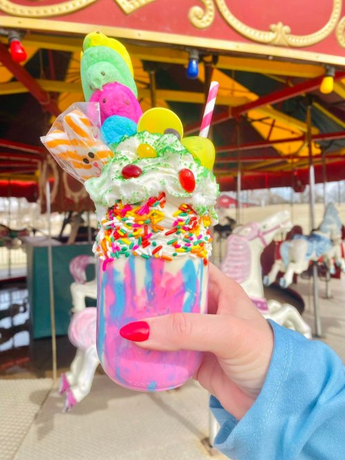 A colorful milkshake topped with whipped cream, sprinkles, and candy, held in front of a carousel.