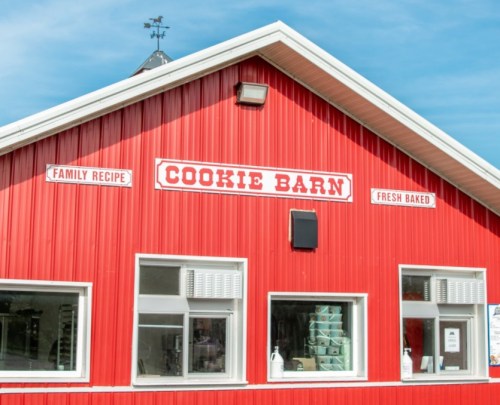 Red barn-style building with signs reading "Cookie Barn," "Family Recipe," and "Fresh Baked." Clear blue sky above.