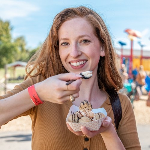 A smiling woman holds a bowl of ice cream with a spoon, enjoying a sunny day at a park.