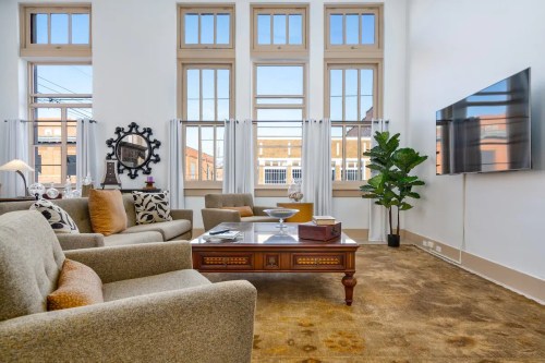 Bright living room with large windows, comfortable seating, a coffee table, and a TV on the wall.