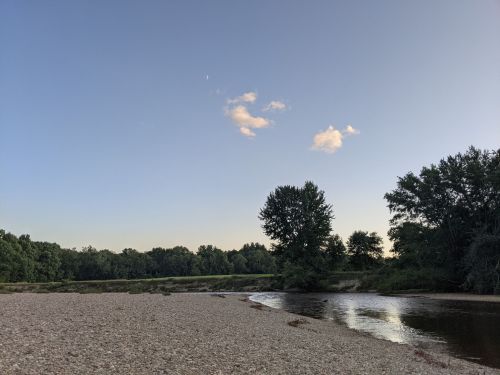 A serene riverbank scene at dusk, featuring a calm river, sandy shore, and trees under a clear sky with a few clouds.