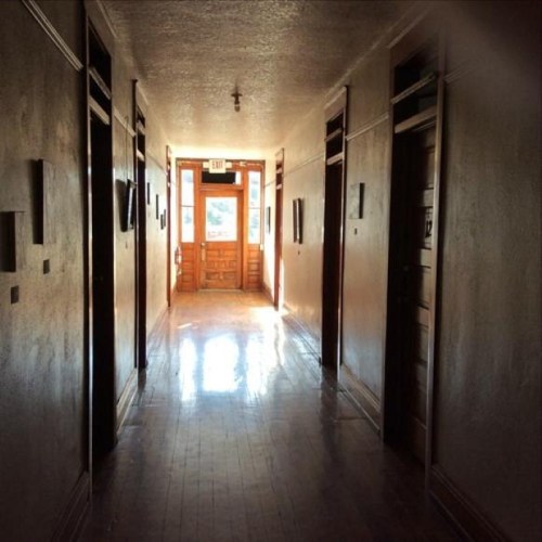 A long, narrow hallway with wooden floors and doors on either side, leading to a bright exit at the end.