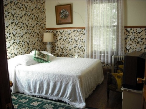 Cozy bedroom with a white bedspread, floral wallpaper, a window with sheer curtains, and a vintage lamp.