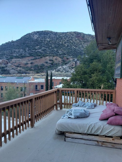 A cozy outdoor balcony with a bed and pillows, overlooking a mountainous landscape and nearby buildings.