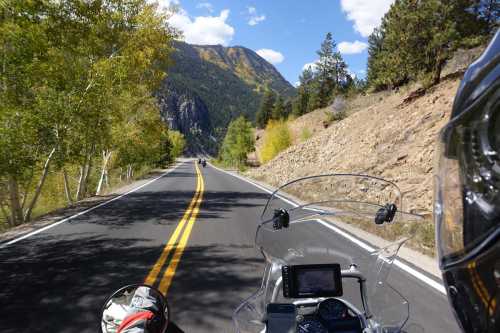 A scenic mountain road with yellow lines, surrounded by trees and rocky terrain, viewed from a motorcycle.