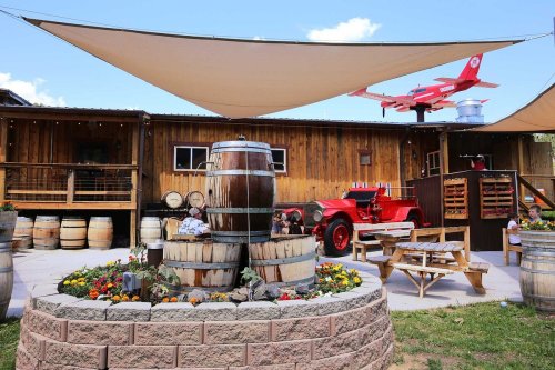 A rustic outdoor space with wooden barrels, a fountain, colorful flowers, and a vintage red plane above a cozy seating area.
