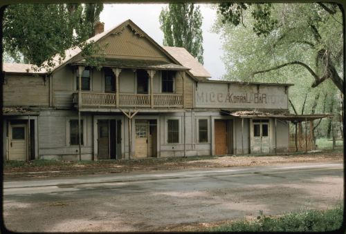 Abandoned building with a porch, overgrown trees, and a faded sign reading "Mice Korral Bar." Road in front.