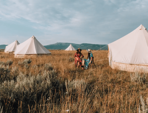Two people in colorful outfits sit near glamping tents in a grassy field, with mountains in the background under a blue sky.