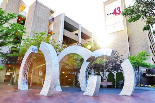 A modern archway sculpture in an urban setting, surrounded by trees and buildings, with sunlight shining through.
