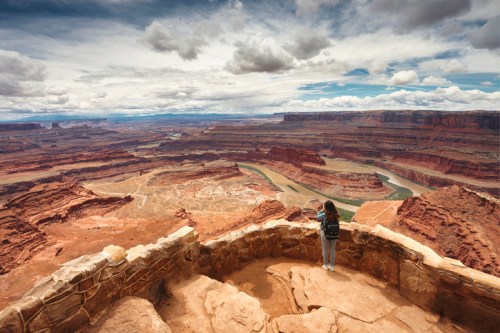 A person stands on a stone overlook, gazing at a vast canyon landscape under a cloudy sky.