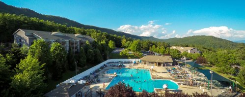 Aerial view of a resort with a large pool, surrounded by trees and mountains under a clear blue sky.