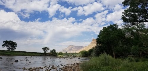 A serene river scene with rocky banks, lush greenery, and a backdrop of mountains under a partly cloudy sky.