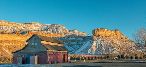 A red barn sits in front of snow-capped mountains under a clear blue sky, showcasing a serene rural landscape.