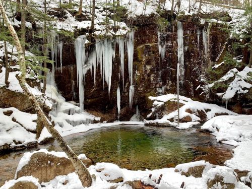 A serene winter scene featuring a rocky wall with icicles, a small green pool, and snow-covered ground.