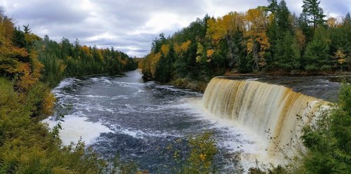 A scenic view of a waterfall surrounded by autumn foliage and a river, under a cloudy sky.
