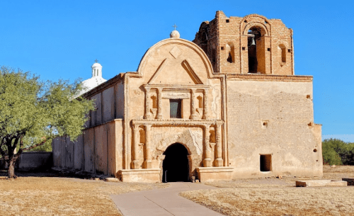 Historic adobe church with a bell tower, surrounded by dry grass and clear blue sky.