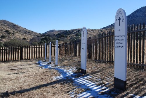 Grave markers in a rural cemetery, with mountains in the background and a wooden fence surrounding the area.