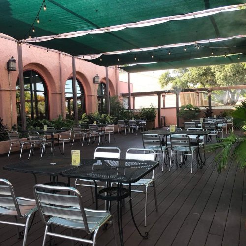 Outdoor dining area with metal tables and chairs, shaded by a green canopy, surrounded by plants and warm-colored walls.