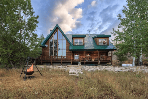 A rustic wooden cabin with a green metal roof, surrounded by trees and a fire pit in the foreground.