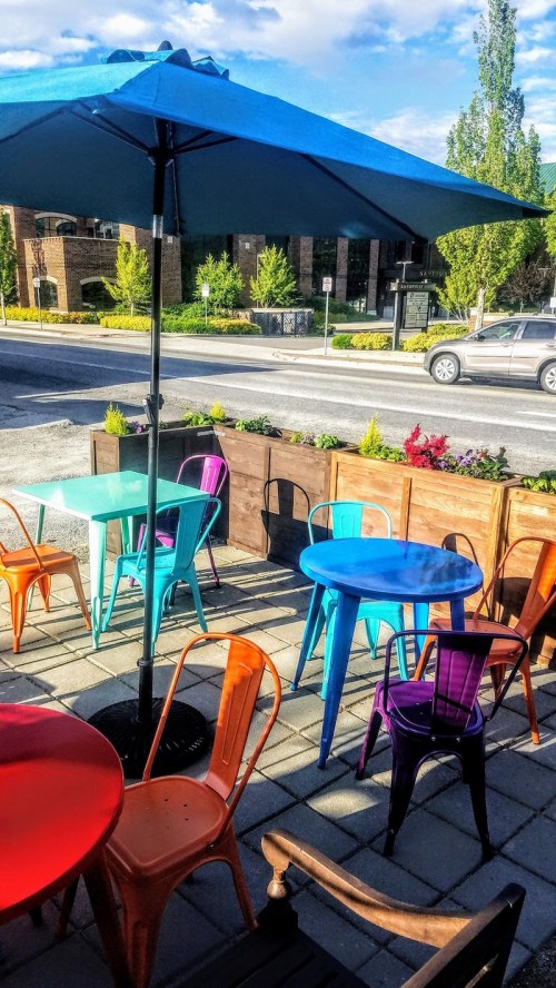 Colorful outdoor seating area with blue umbrellas, vibrant chairs, and a view of a street lined with greenery.