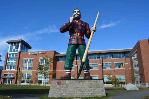 A large statue of a lumberjack in plaid holding a log stands in front of a modern building under a clear blue sky.