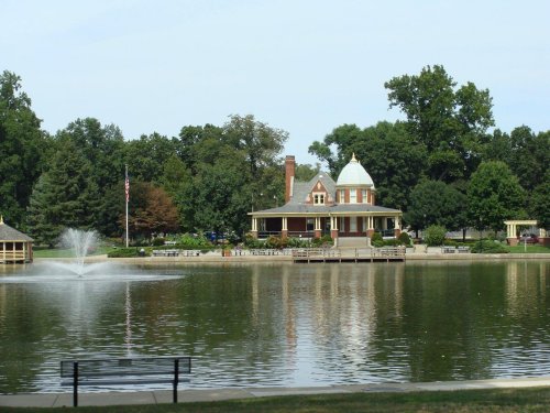 A serene lake scene featuring a gazebo, fountain, and a historic house surrounded by trees.