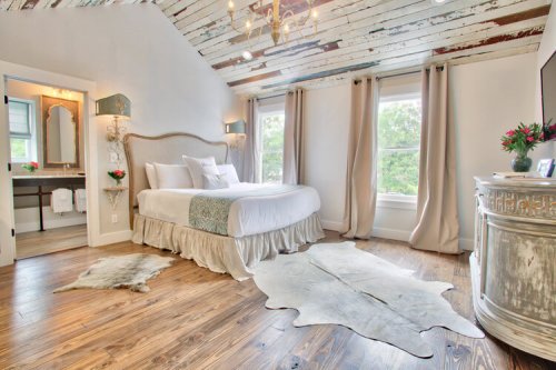 Bright bedroom with a large bed, rustic ceiling, wooden floors, and a cozy rug, featuring natural light from large windows.