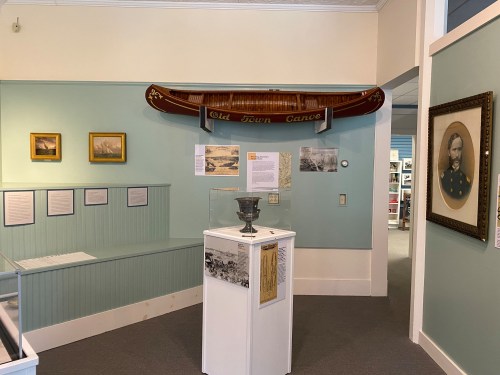 A museum interior featuring a canoe display, framed artworks, and a historical portrait on light green walls.